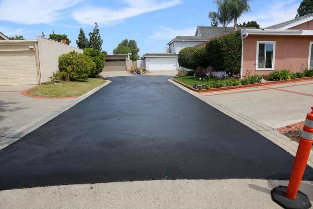 A freshly paved asphalt driveway leading to two garages, framed by grass and shrubbery. On the right, there's a pink house with a well-kept garden. An orange traffic cone is visible on the pavement at the bottom right.