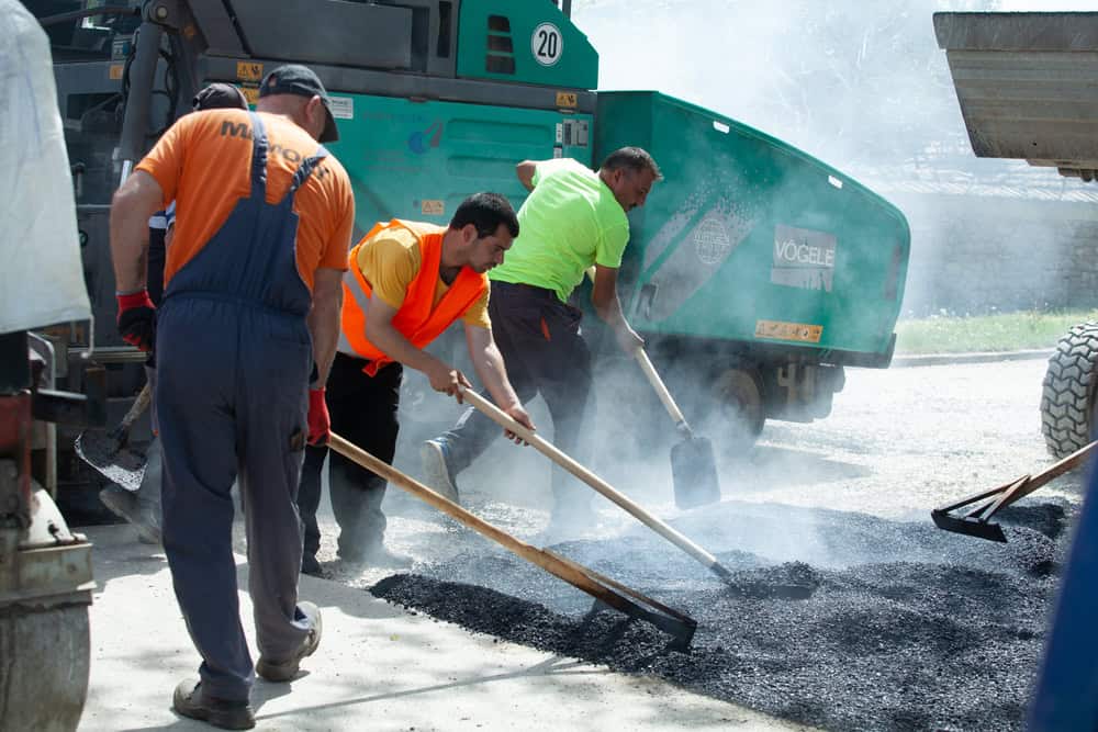 Three construction workers are spreading hot asphalt on a road. They are using wooden-handled rakes and wearing brightly colored safety vests. Smoke rises from the heated material. In the background is roadwork equipment with wheels and a green body.