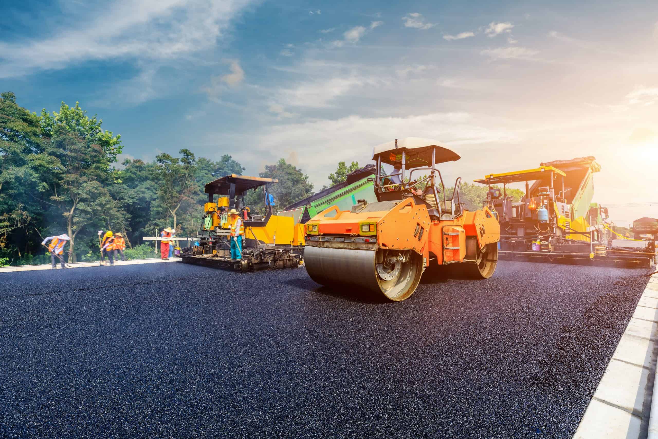 Road construction scene with workers and machinery. An asphalt roller is in the foreground, compacting new black pavement. Behind, two large paving machines operate with workers in helmets and vests. Trees and a clear sky are in the background.
