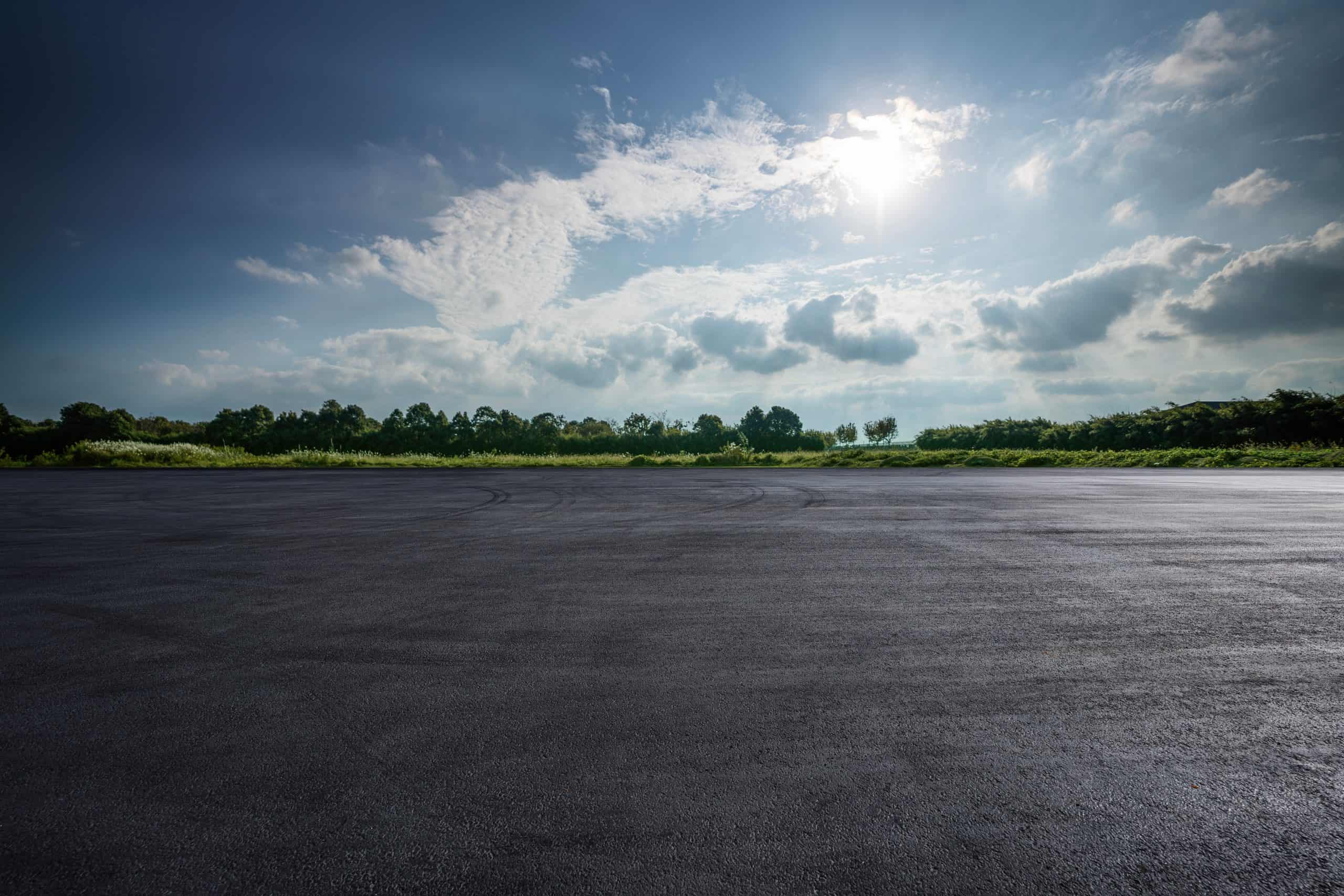 A wide, freshly paved asphalt surface stretches out under a partly cloudy sky. Sunlight breaks through the clouds, illuminating the green treeline in the distance.