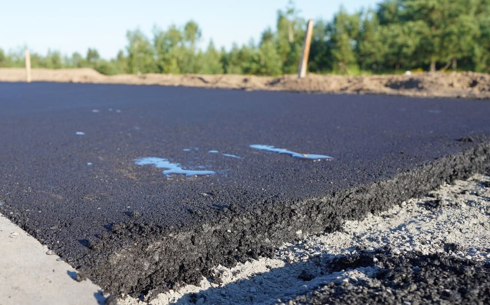 Close-up of fresh asphalt laid on a road, with a visible cross-section showing layered structure. A few patches of water glisten on the surface under clear blue sky. Trees and a dirt embankment are in the background.