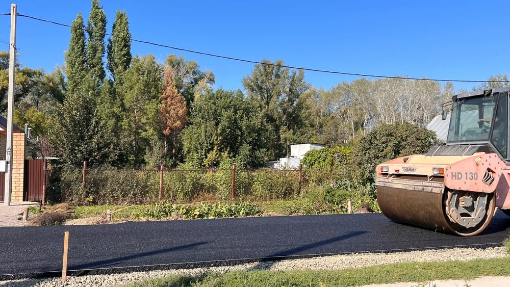 A steamroller smooths freshly laid asphalt on a road adjacent to a grassy area and trees under a clear blue sky. A brick building stands on the left, and power lines stretch across the scene.