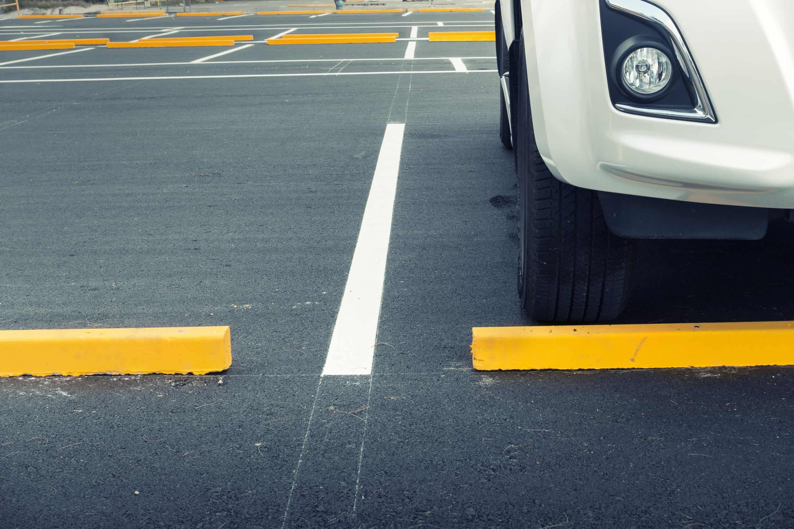 White car partially visible, parked in an empty asphalt parking lot with yellow wheel stops. The car’s right front headlight is shown, facing towards the stops. White lines mark the parking spaces.