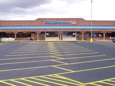 An empty parking lot with yellow lines in front of a brick grocery store, featuring a blue awning and a cloudy sky in the background—a fine example of expert parking lot paving by contractors from Hillsborough County, NH.